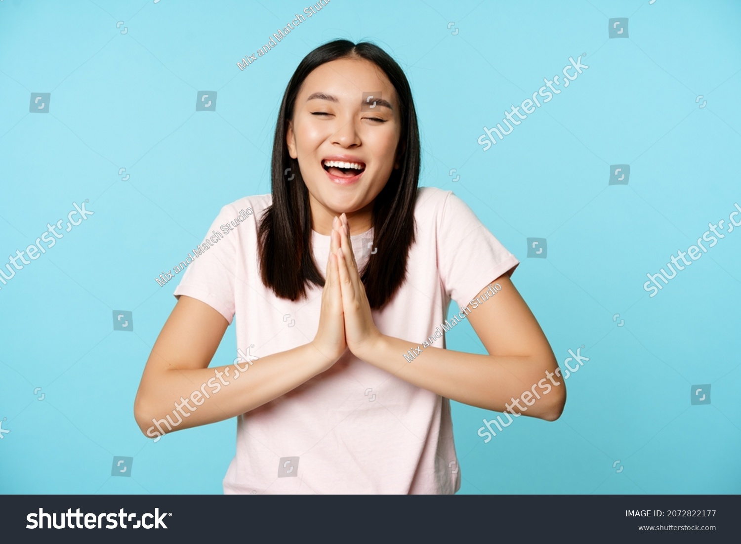 Smiling happy asian woman shows namaste, beg gesture and dreaming of something, making wish, high hopes, standing over blue background #2072822177