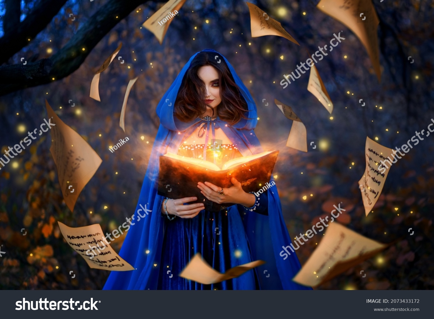 Fantasy woman witch magician in hood holds in hands magic book, bright orange light spells, wind scatters fall sheets paper page levitation. Girl sorceress. Medieval cloak blue dress magician costume #2073433172