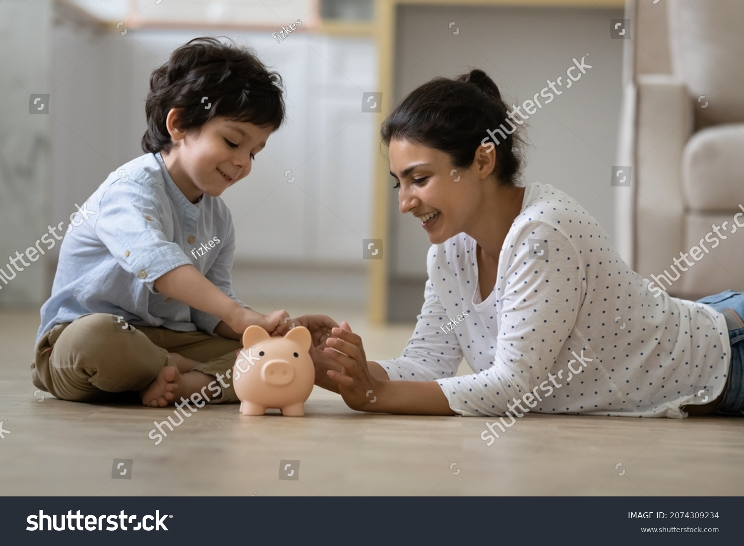 Happy Indian kid and young mom saving money together, putting cash into ceramic piggy bank. Mother playing with child on heating floor at home, teaching little son to invest money, planning future #2074309234