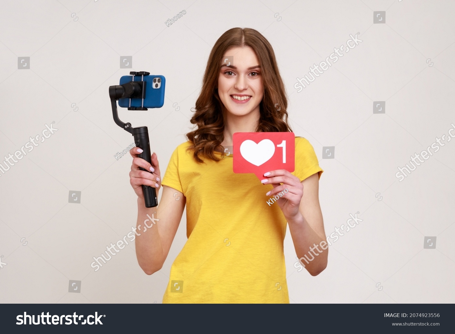 Smiling teenage girl shoots herself on electronic stabilizer for video shooting on phone, broadcasting livestream, showing like icon. Indoor studio shot isolated on gray background. #2074923556