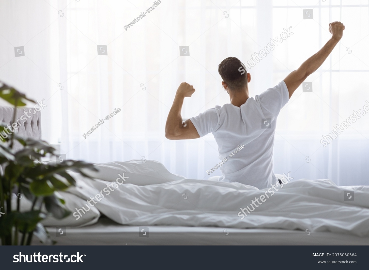 Rear View Of Young Man Stretching In Bed After Waking Up In The Morning, Unrecognizable Male Resting In Light Bedroom After Good Sleep, Looking At Window, Enjoying Start Of New Day, Copy Space #2075050564