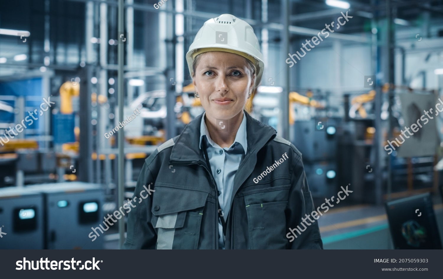 Car Factory Office: Portrait of Female Chief Engineer Wearing Hard Hat Looking at Camera, Smiling. Professional Technician. Automated Robot Arm Assembly Line Manufacturing High-Tech Electric Vehicles #2075059303