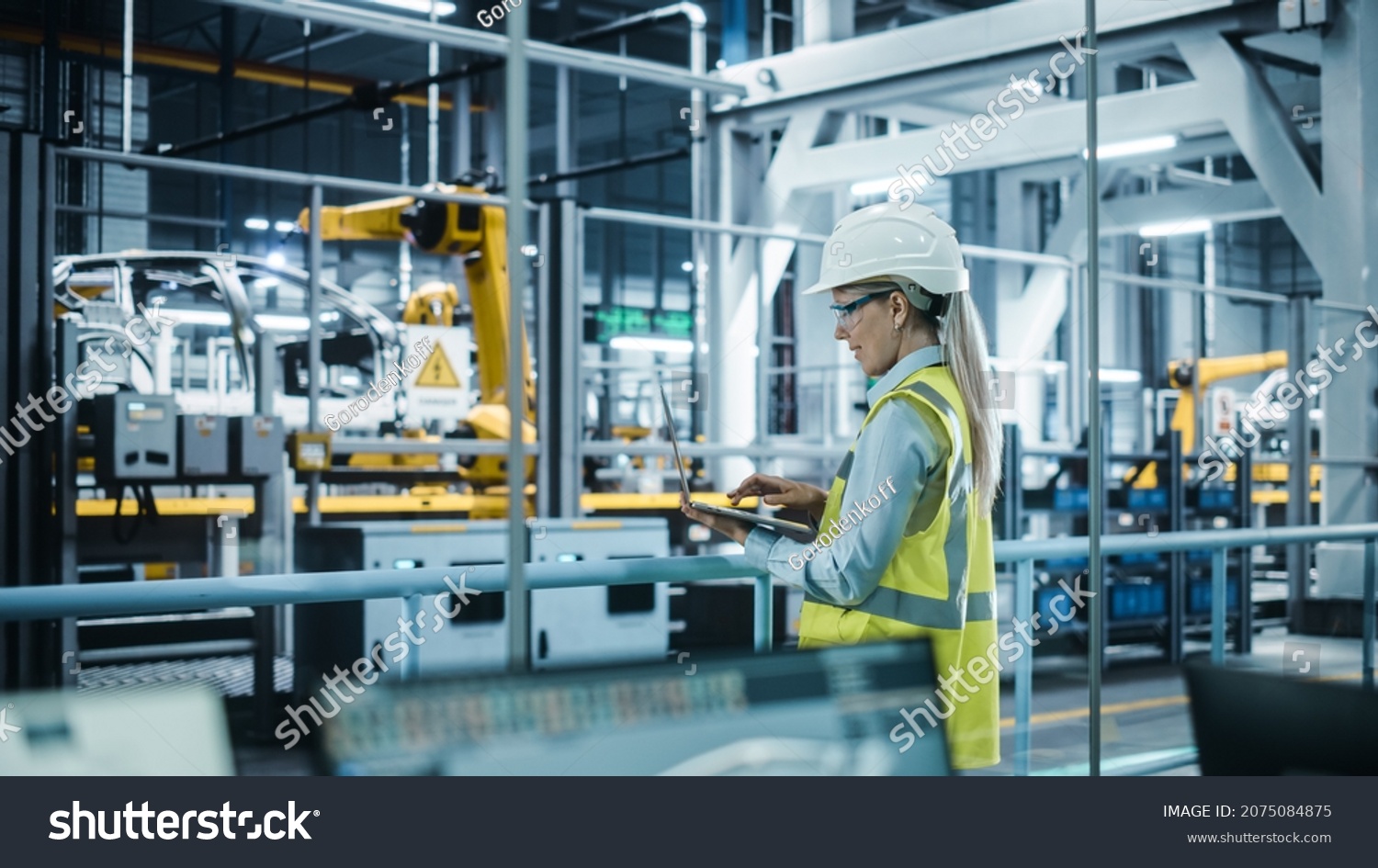 Car Factory: Female Automotive Engineer Wearing Hard Hat, Standing, Using Laptop. Monitoring, Control, Equipment Production. Automated Robot Arm Assembly Line Manufacturing Electric Vehicles. #2075084875