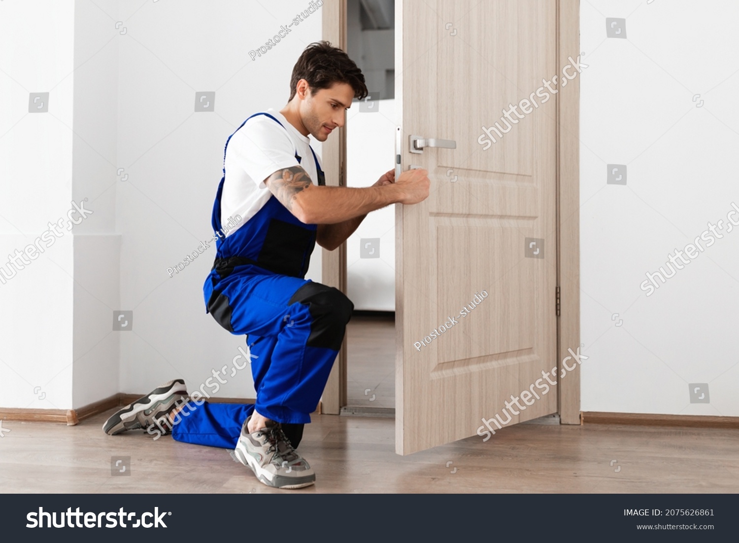 Installation of a lock on the front wooden entrance door. Portrait of young locksmith workman in blue uniform installing door knob. Professional repair service. Maintenance Concept #2075626861