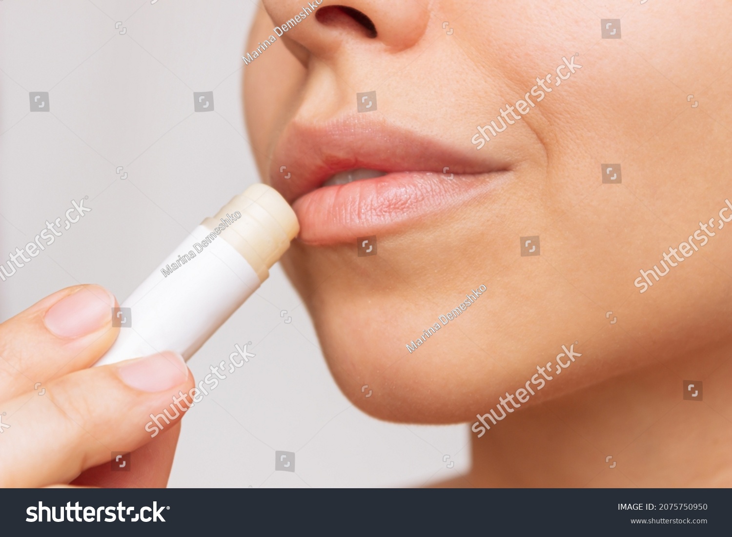 Cropped shot of a young caucasian beautiful woman applying a hygienic lipstick on her lips on a gray background. Moisturizing chapstick for dry lips #2075750950