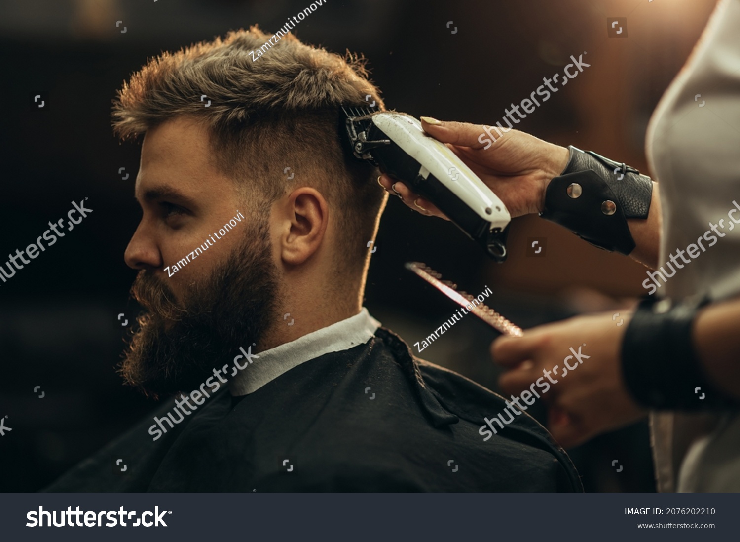 Young bearded man getting haircut by hairdresser while sitting in chair at barbershop #2076202210