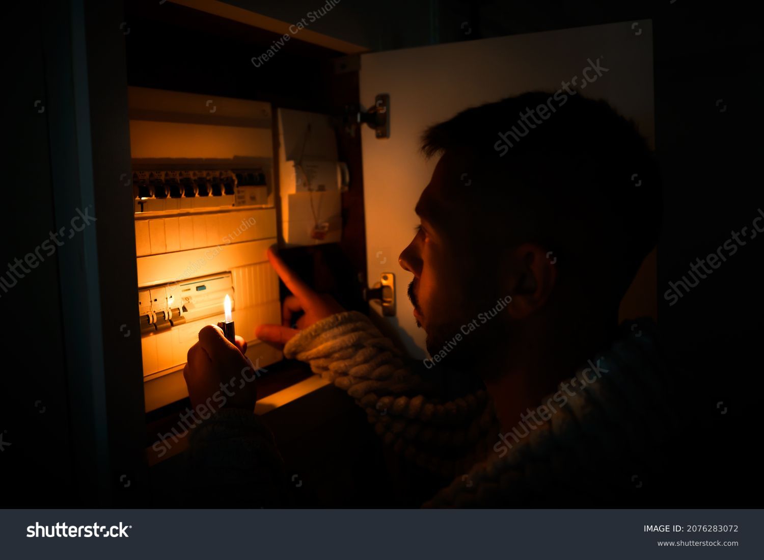 Man with lighter in total darkness investigating fuse box or electric switchboard at home during power outage. Blackout, no electricity concept #2076283072