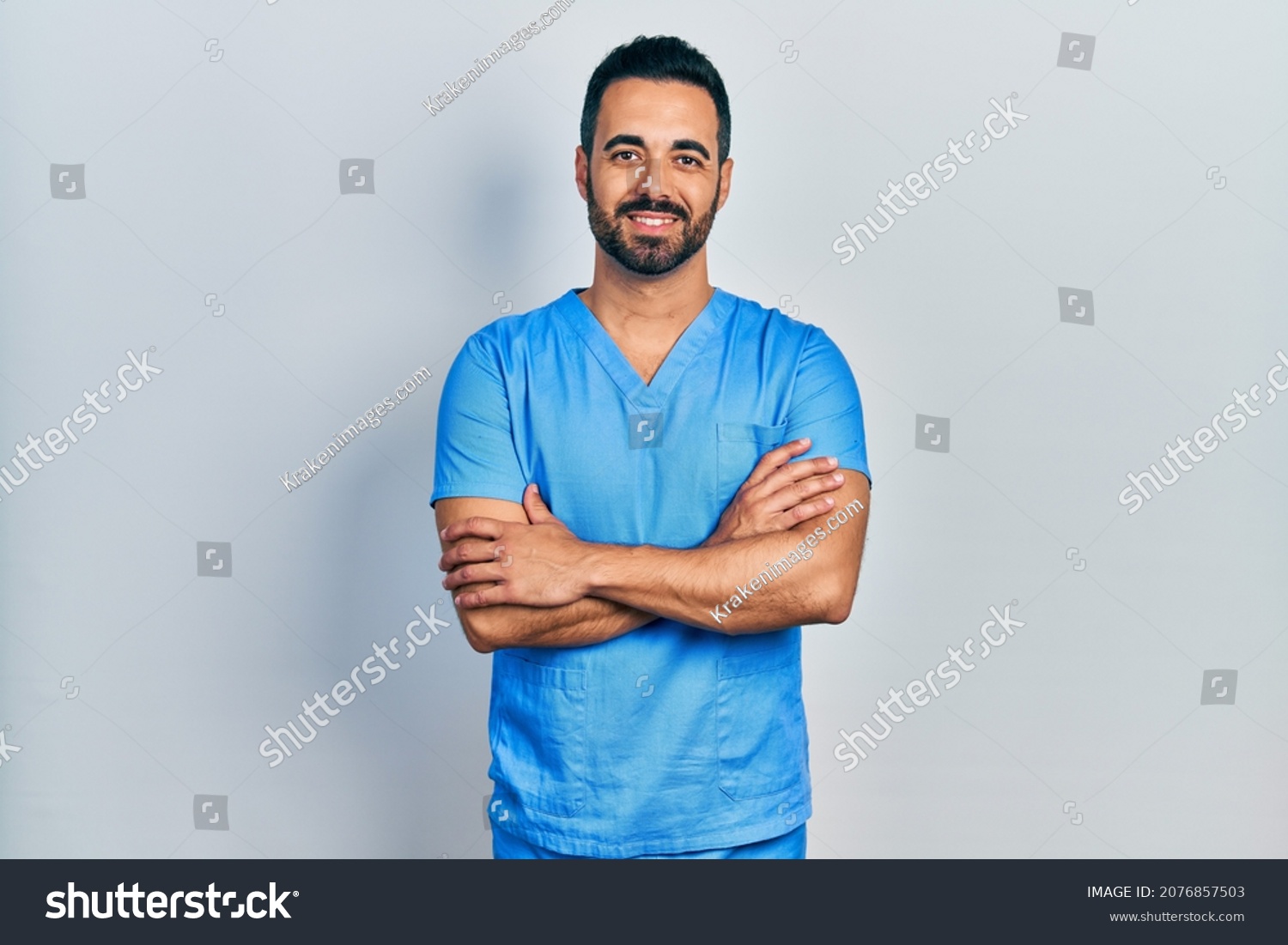 Handsome hispanic man with beard wearing blue male nurse uniform happy face smiling with crossed arms looking at the camera. positive person.  #2076857503