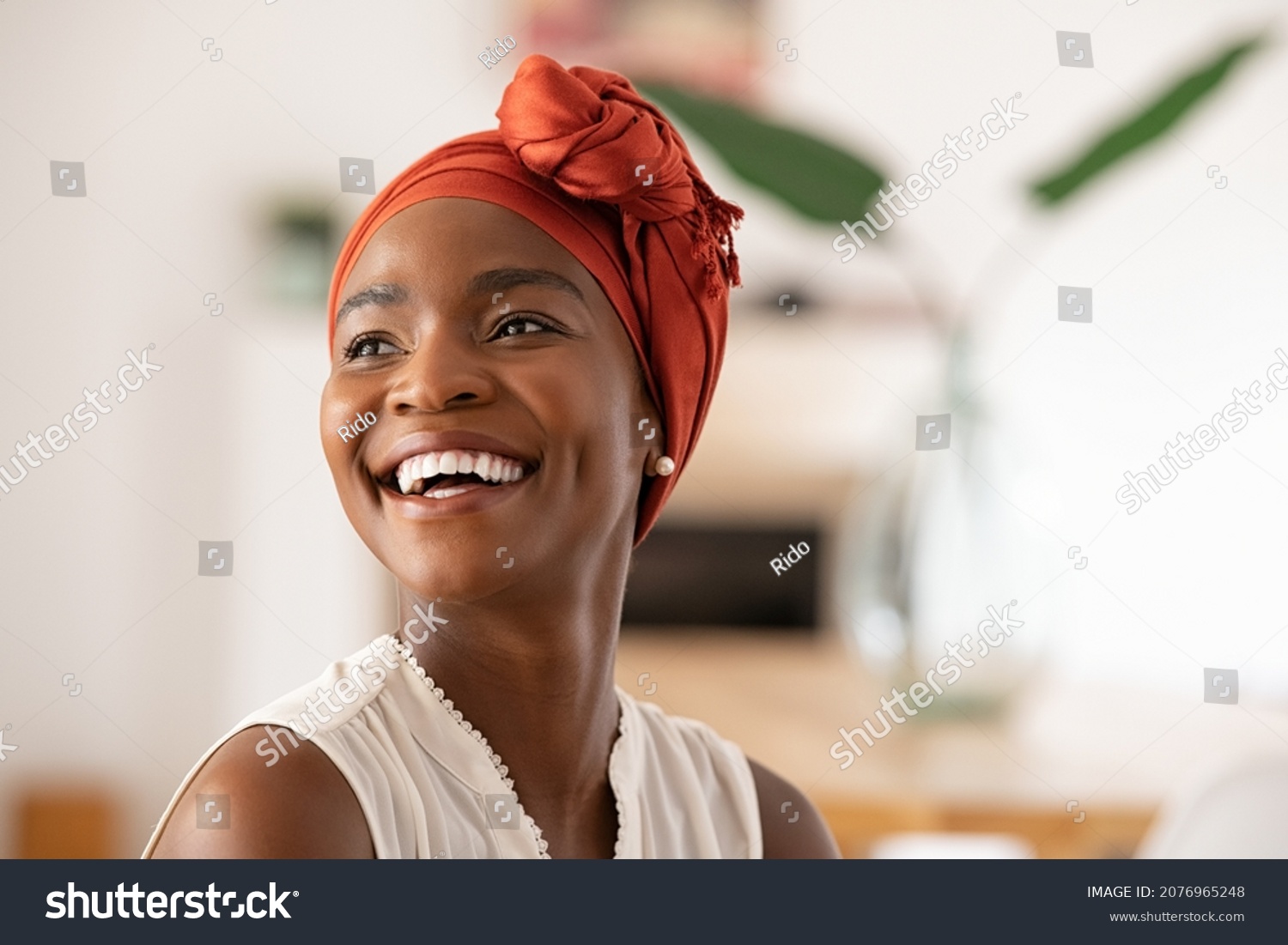 Beautiful mature black woman with african turban relaxing at home. Cheerful middle aged black woman in casual clothing with traditional headscarf at home laughing. African american lady smiling. #2076965248