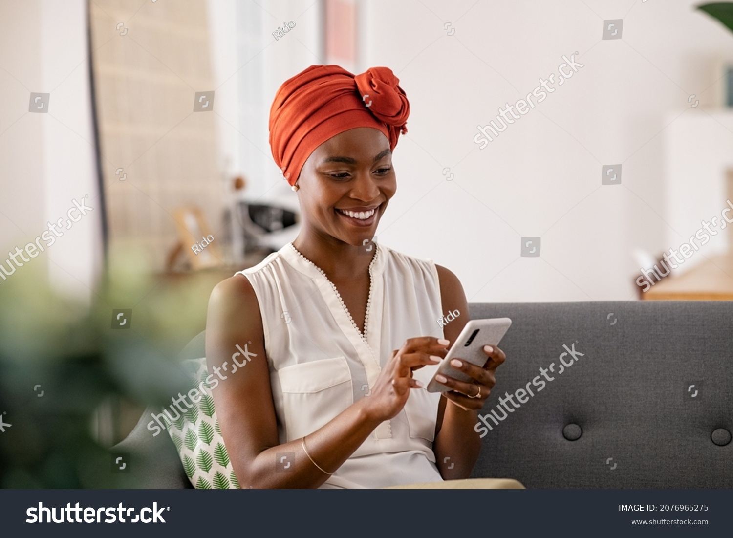 Smiling middle aged african woman with traditional head turban sitting on couch at home using smartphone. Beautiful african american woman with typical headscarf scrolling through internet on phone. #2076965275