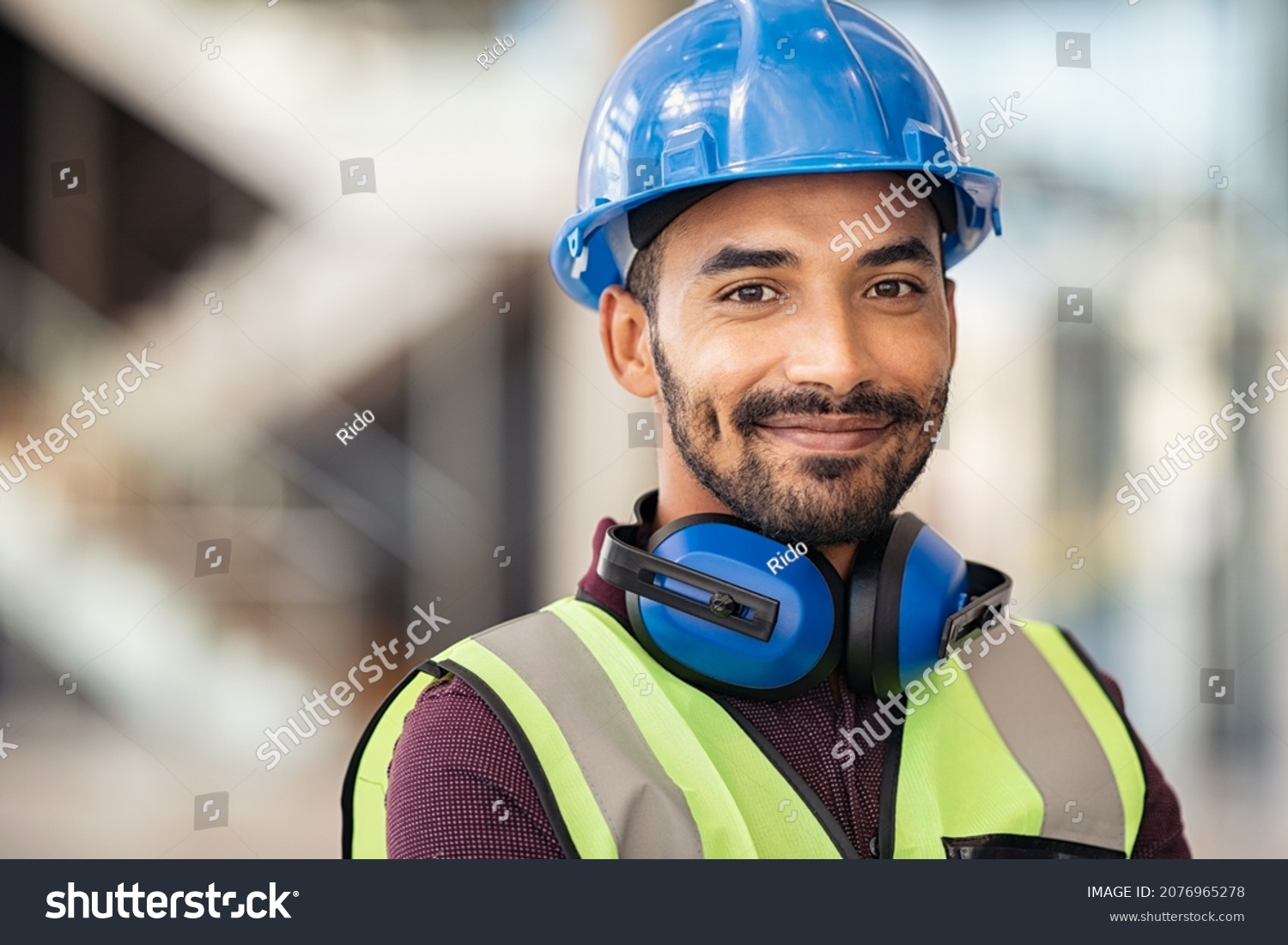 Portrait of satisfied construction site manager wearing safety vest and blue helmet with copy space. Young middle eastern architect watching construction site with confidence and looking at camera. #2076965278