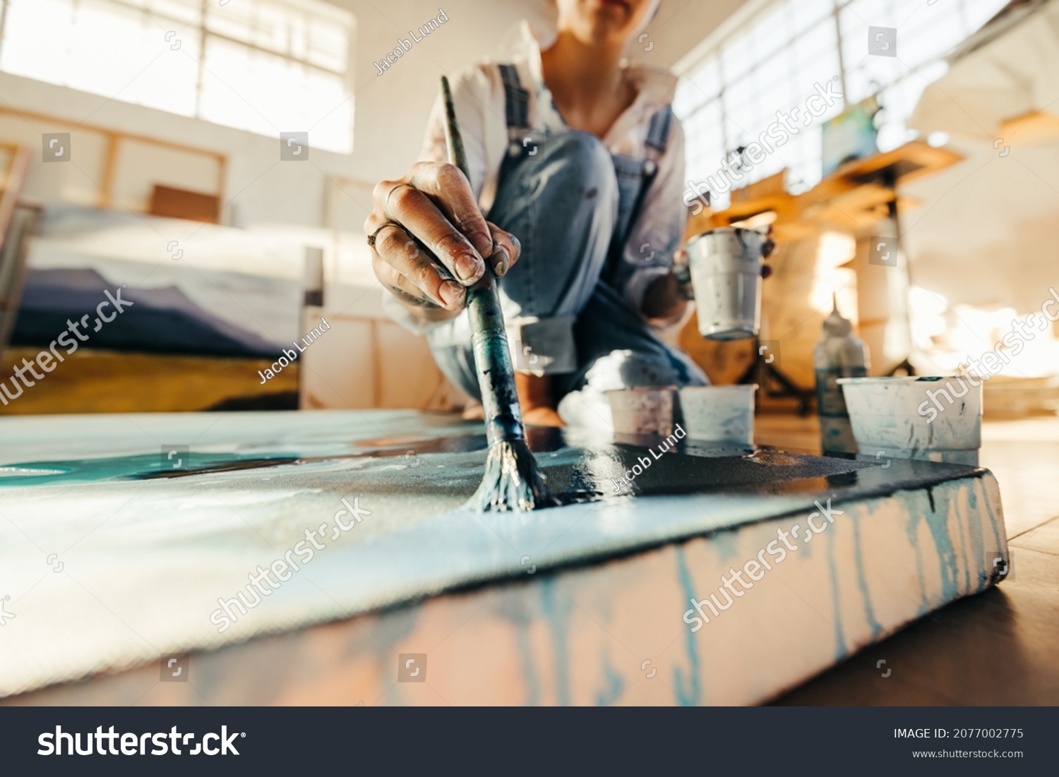 Unrecognizable female artist painting on a large canvas with a paintbrush. Young painter working on the floor of her art studio. Creative young woman making a blue painting for her new art project. #2077002775