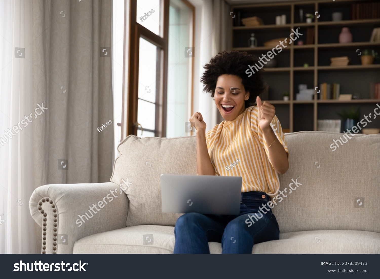 Excited cheerful young Black woman using laptop computer on sofa at home, getting good news, feeling joy, dancing with hands, singing, laughing, making winner gesture, happy to win prize #2078309473
