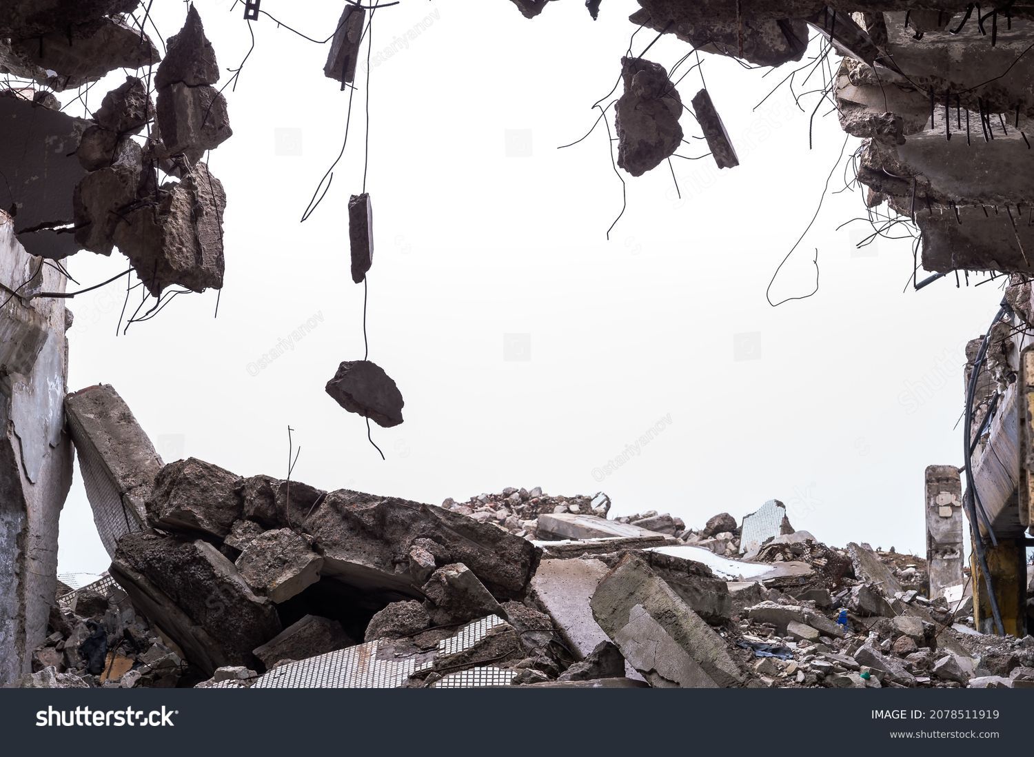 A hole in the body of a building with a pile of construction debris and concrete fragments hanging on the rebar against a uniform gray sky. Background. #2078511919
