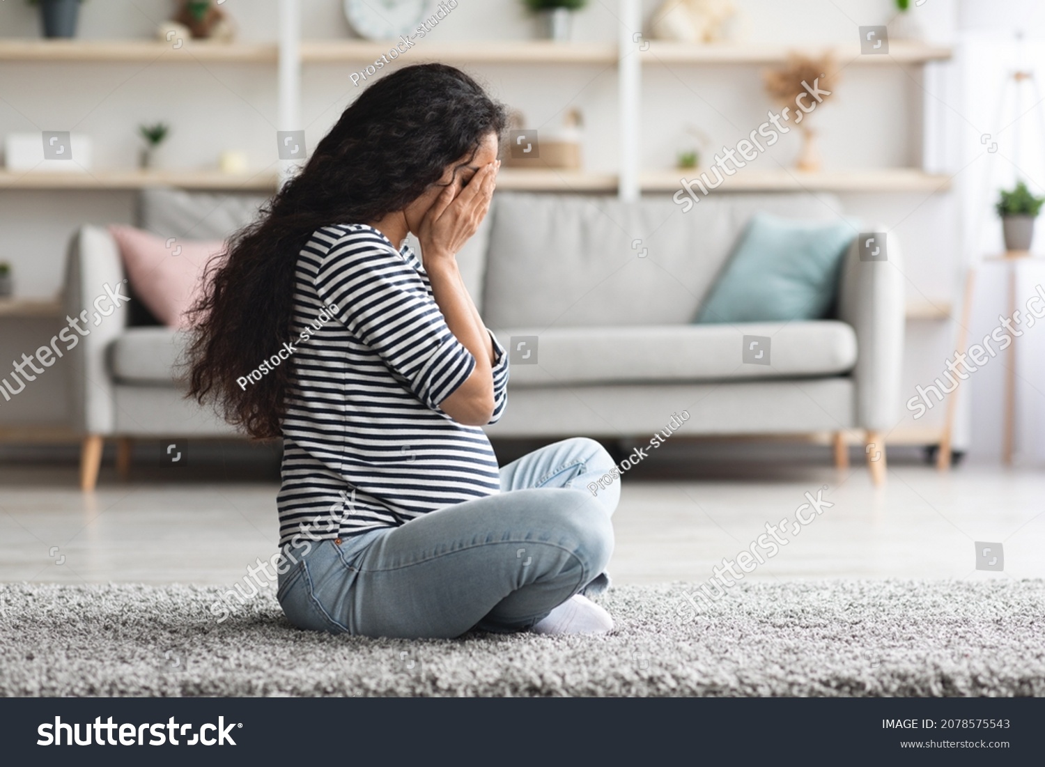 Pregnancy hormonal changes concept. Young curly long-haired pregnant woman feeling down, sitting on floor in cozy living room and crying, staying alone at home, side view, free space #2078575543