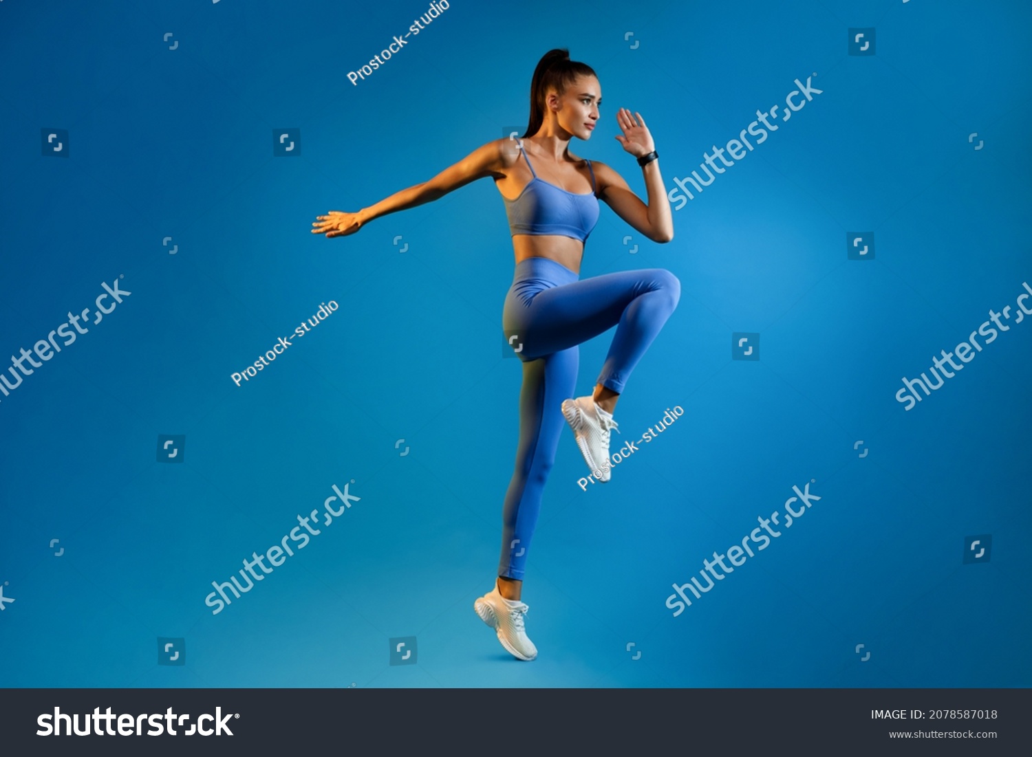 Female Athlete Jumping Exercising During Training Over Blue Studio Background, Looking Aside. Fitness Workout And Sport Motivation Concept. Full Length, Side View #2078587018