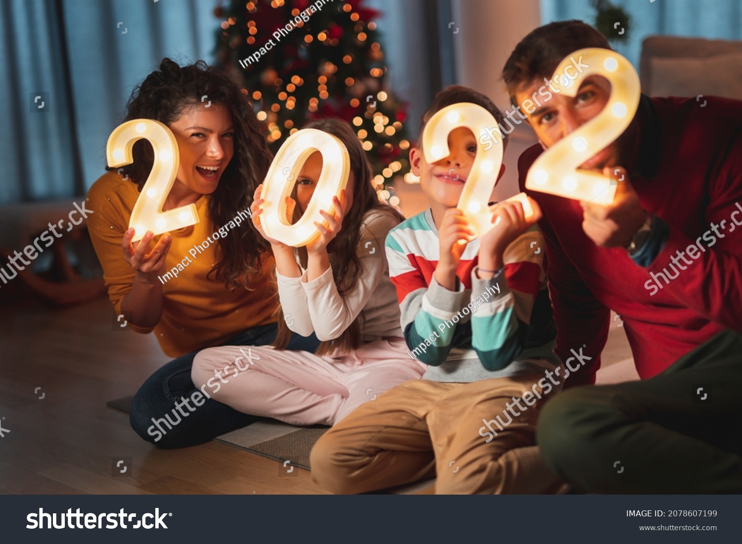 Happy family celebrating New Years Eve at home with kids, sitting by the Christmas tree, holding  illuminative numbers 2022 representing the upcoming New Year #2078607199
