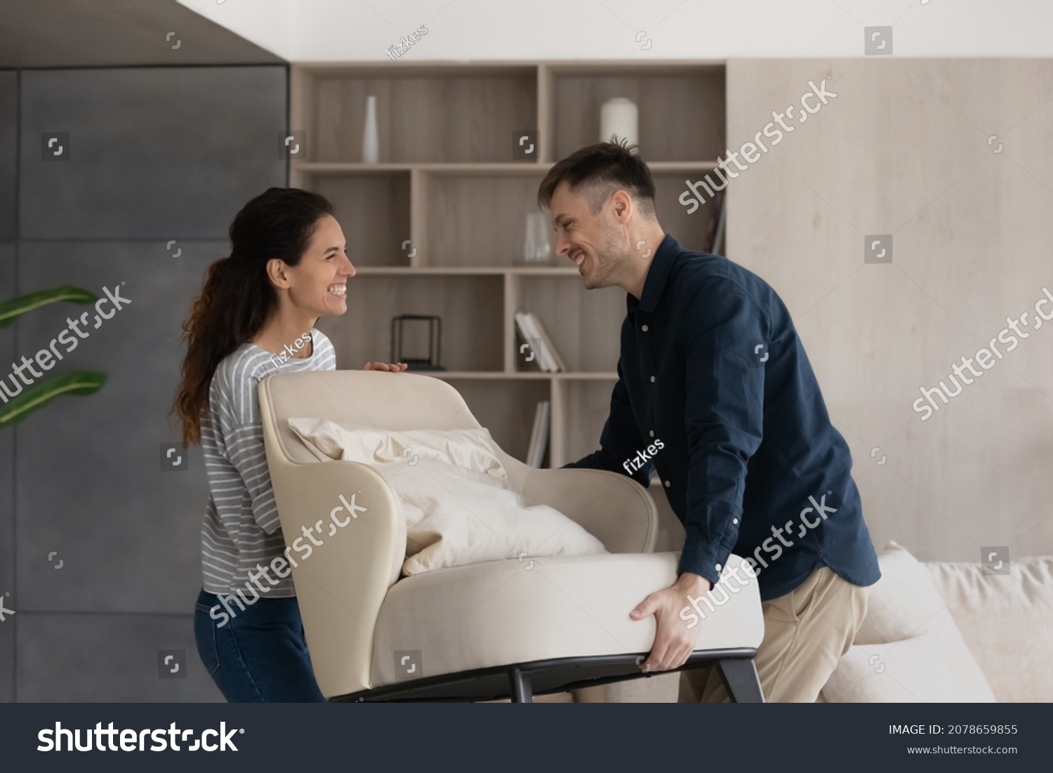 Moving day, happy buyers of fashionable furniture, bank mortgage concept. Smiling loving millennial wife and husband carry new armchair into living room, making home design improvements feel satisfied #2078659855