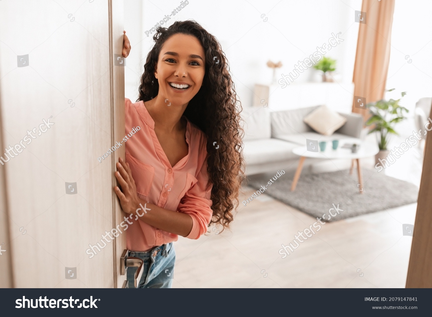Welcome. Portrait of cheerful woman standing in doorway of modern apartment, greeting visitor and inviting guest to enter her home, happy smiling young lady holding door looking out flat #2079147841