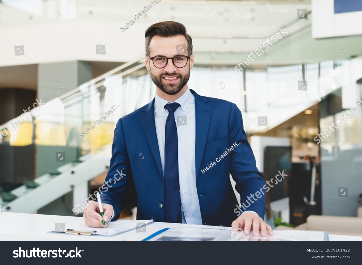 Successful caucasian smiling man shop assistant receptionist in formal attire writing looking at camera while standing at reception desk in hotel car dealer shop #2079161422