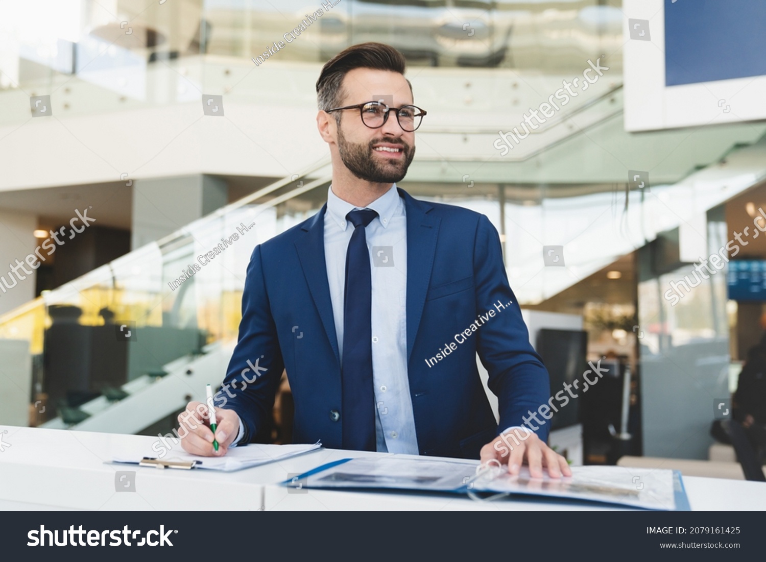 Successful caucasian smiling man shop assistant receptionist in formal attire writing while standing at reception desk in hotel car dealer shop #2079161425