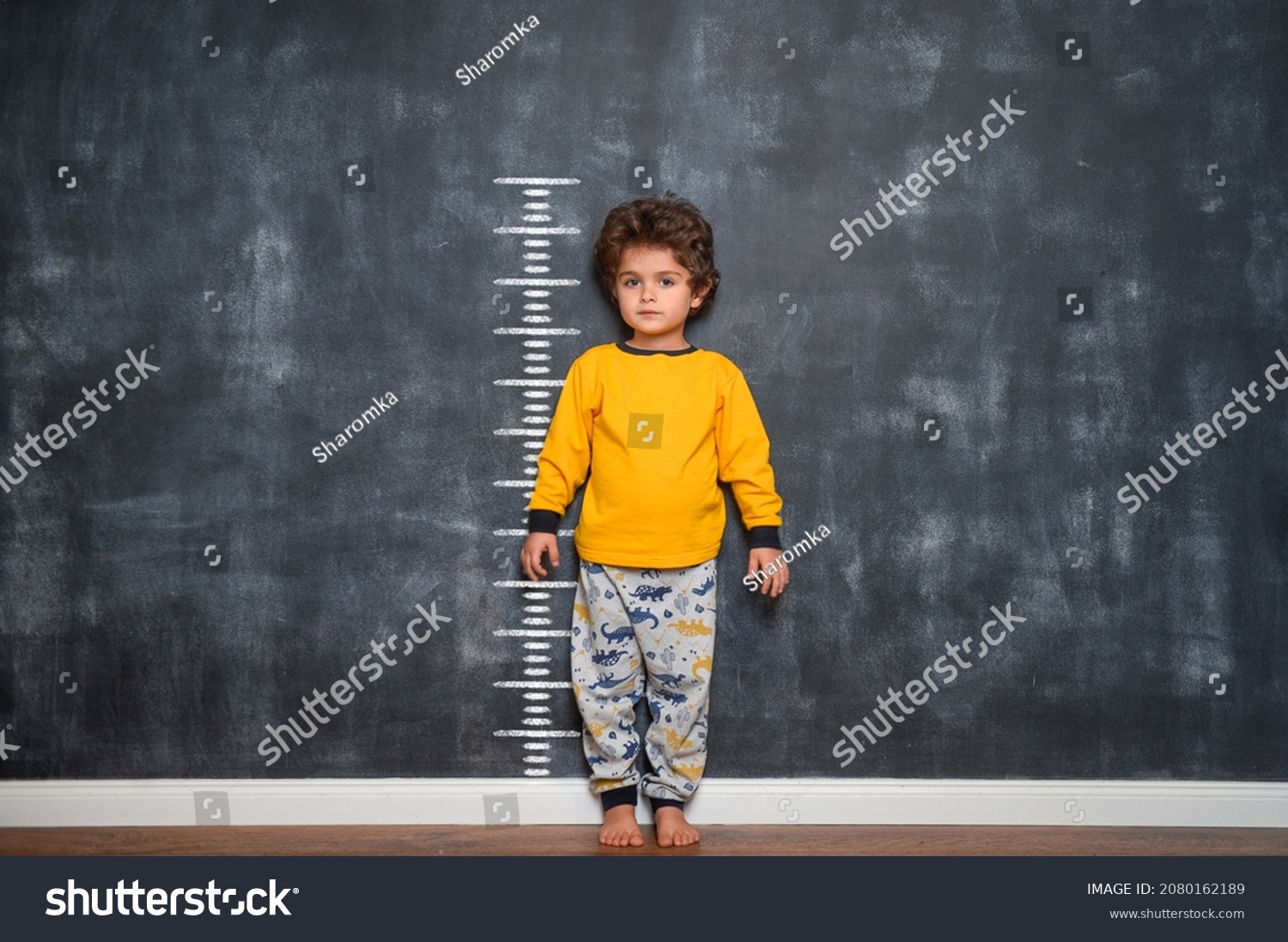 Funny smiling little child with measures of growth on blackboard wall beside him. Confident kid from kindergarten is ready to go to school. #2080162189