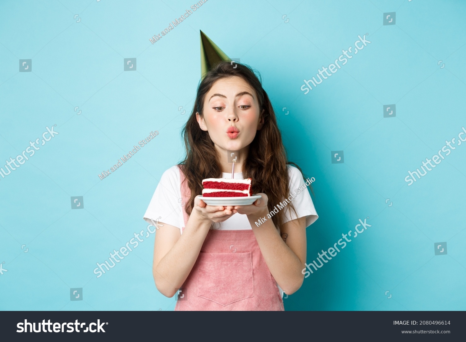 Holidays and celebration. Excited woman celebrating birthday, blowing candle on cake, wearing party cake and having fun, standing over blue background #2080496614