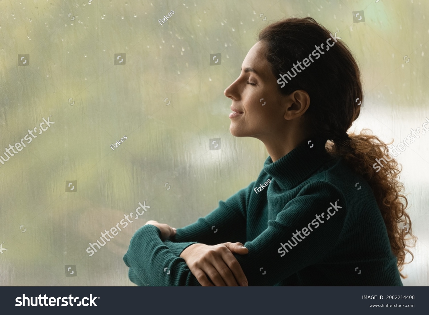 Music of rain. Side shot of serene latin woman relax by window with closed eyes listen sound of raindrops feel pleasure. Calm young lady enjoy breathing fresh ozonized air after rainstorm. Copy space. #2082214408