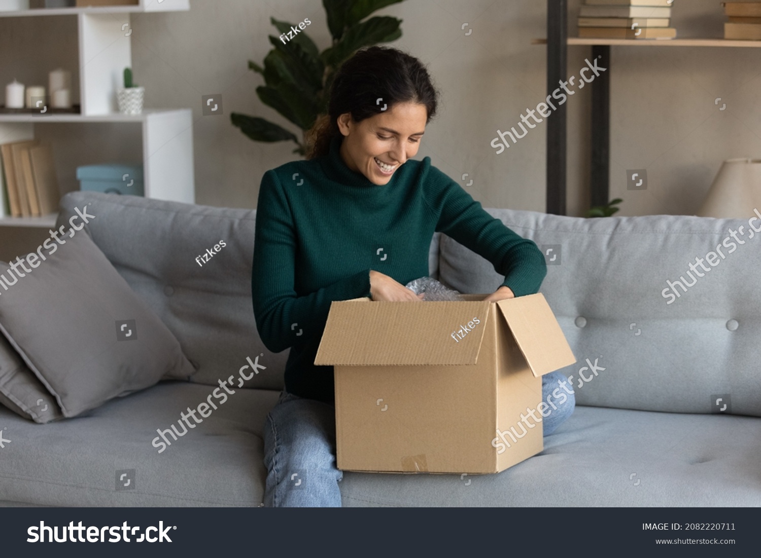 Ecommerce. Satisfied young latin woman web shop customer sit on sofa open postal delivery package with consumer goods products ordered online. Happy millennial lady unpacking after moving to new home #2082220711