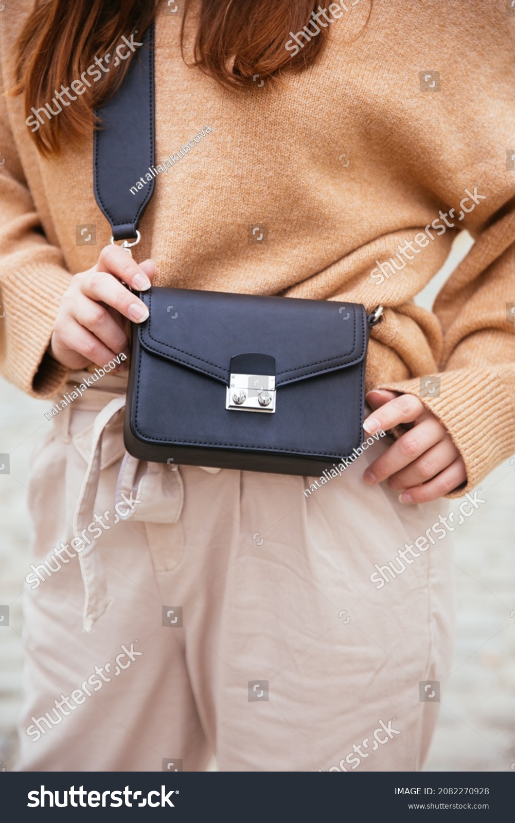 Closeup of black small handbag in woman's hand. Fall spring fashion outfit camel sweater and trendy white jeans. Fashion detail, stylish small bag.   #2082270928
