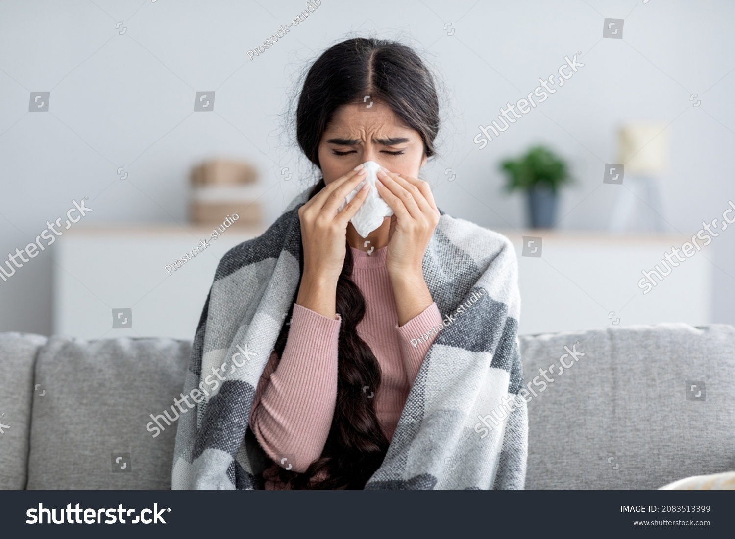 Unhappy sad young indian female in plaid suffering from fever and flu on sofa, blowing nose in napkin in living room interior. Covid-19 lockdown, treatment of illness, cold and runny, copy space #2083513399