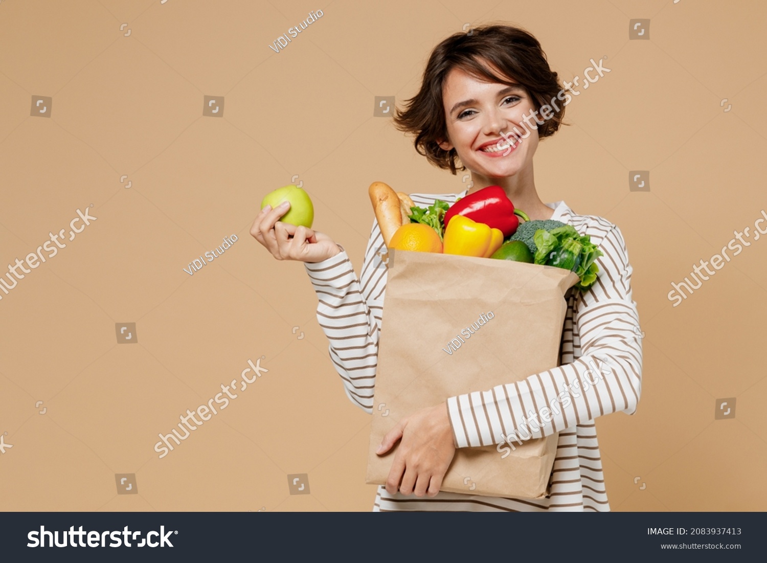 Young smiling vegetarian cheerful woman 20s in casual clothes hold paper bag with vegetables holding apple fruit eating isolated on plain pastel beige background studio portrait. Shopping concept. #2083937413