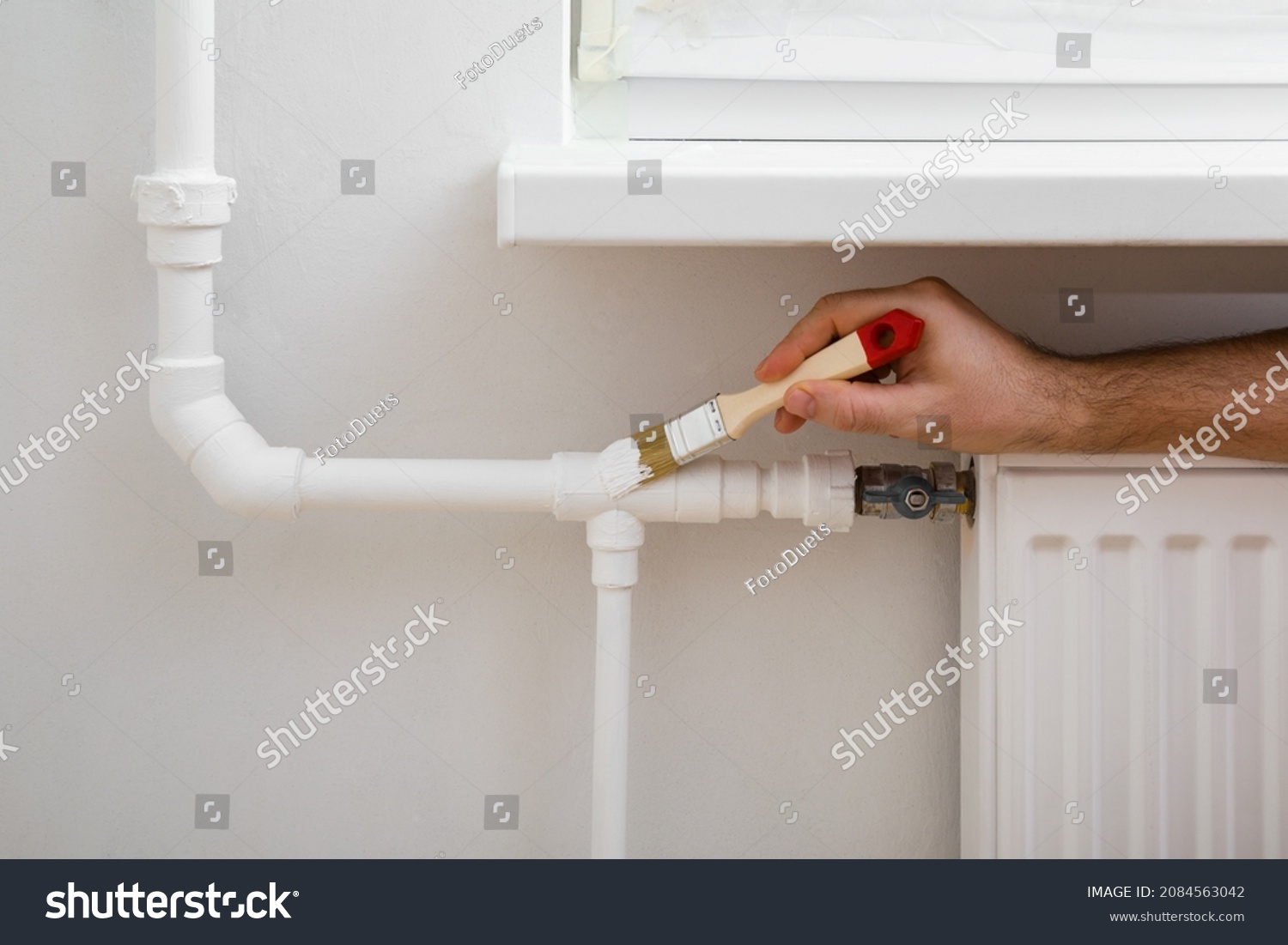 Young adult man hands using brush and painting heating pipe white color. Closeup. Repair work of home. #2084563042