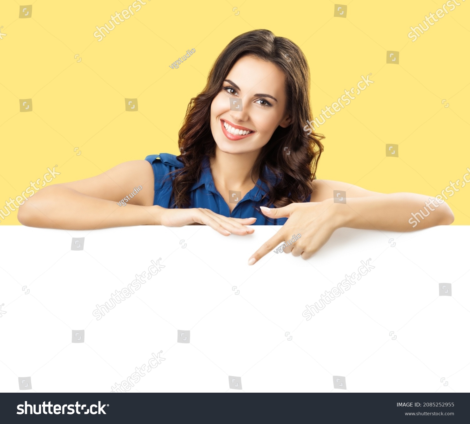 Happy smiling brunette woman in blue dress, showing, pointing, standing behind, peeping from blank banner mock up white billboard sign board signboard with ad copy space, over yellow color background. #2085252955