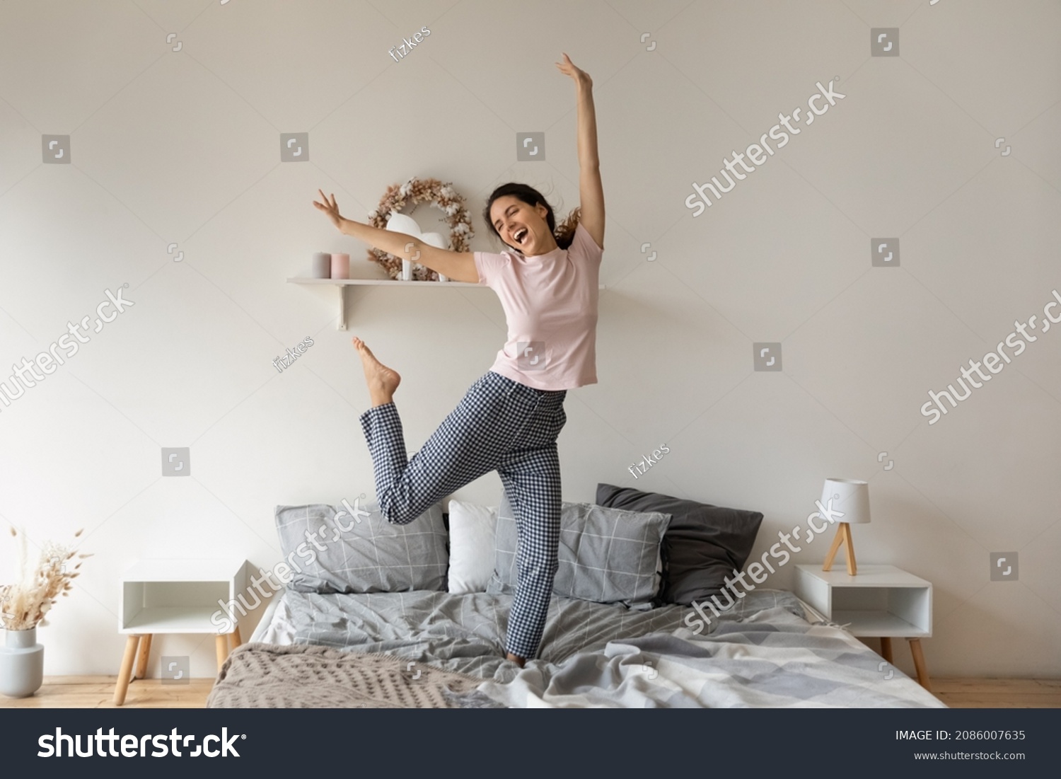 Overjoyed funny woman jumping on comfortable cozy bed at home, enjoying morning, starting new day, excited happy young female wearing pajama dancing to favorite music, having fun in modern bedroom #2086007635