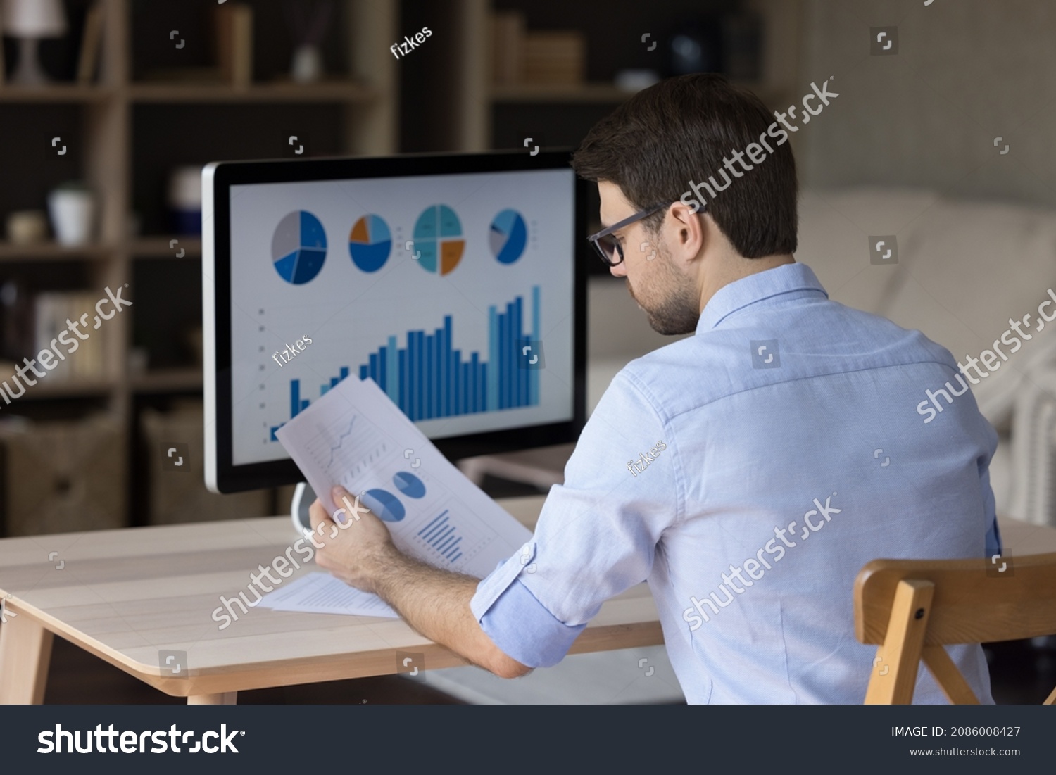 Focused millennial business man analyzing marketing reports on desktop monitor, reviewing paper graphs, financial stats, startup project infographics. Marketer working from home office. Back view #2086008427