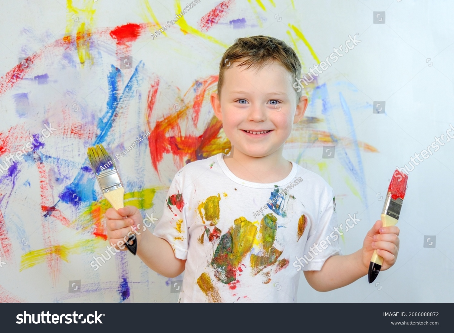 A handsome smiling blue-eyed boy stands against the background of a colorful wall. The child holds a brush in his hands. Boy in a white T-shirt, stained with paint. Wall in colored paints. #2086088872