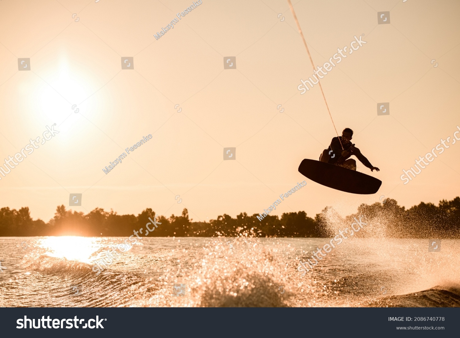 Great view of dark silhouette of active male rider holding rope and making extreme jump on wakeboard over splashing water. Wakeboarding and water sports activity. #2086740778