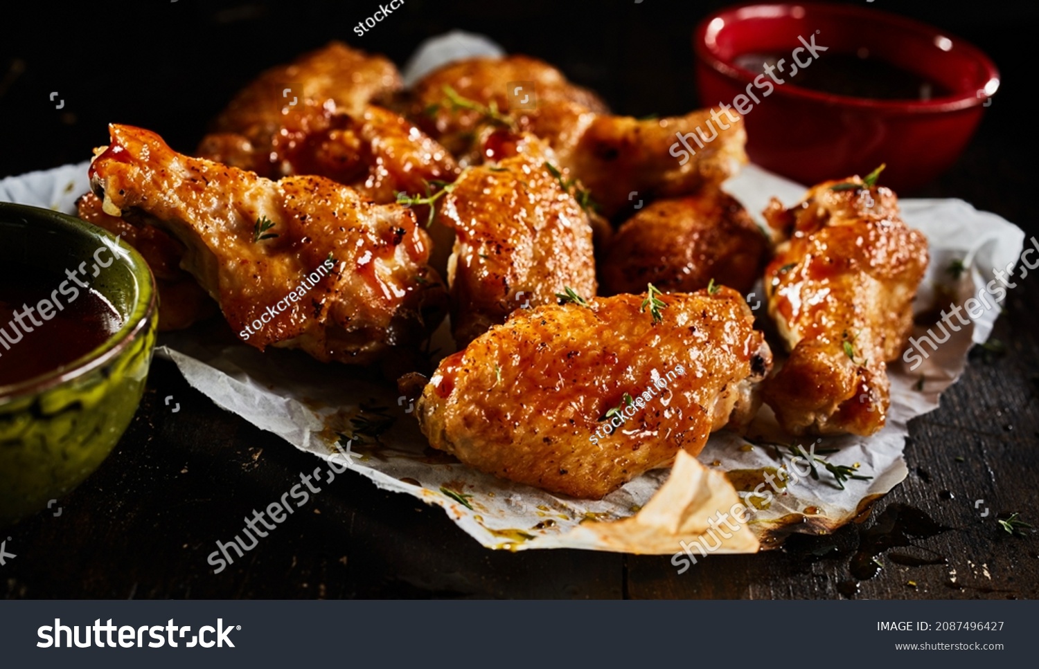 High angle of appetizing BBQ chicken wings with herbs served on paper on dark table with sauce in bowls in studio #2087496427