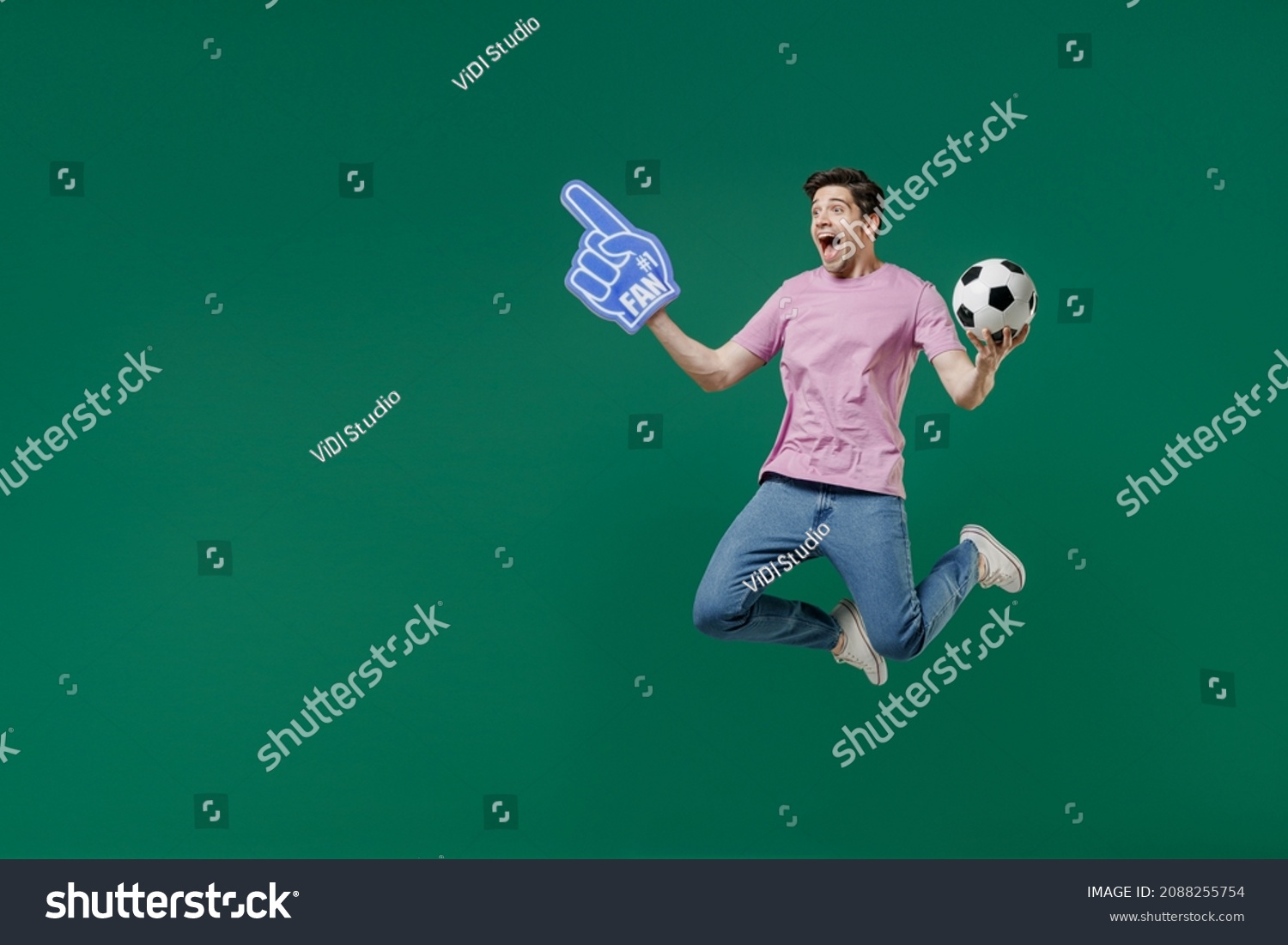 Full size body length young fun man fan wear pink t-shirt cheer up support football sport team hold in hand soccer ball watch tv live stream scream jump isolated on dark green color background studio #2088255754