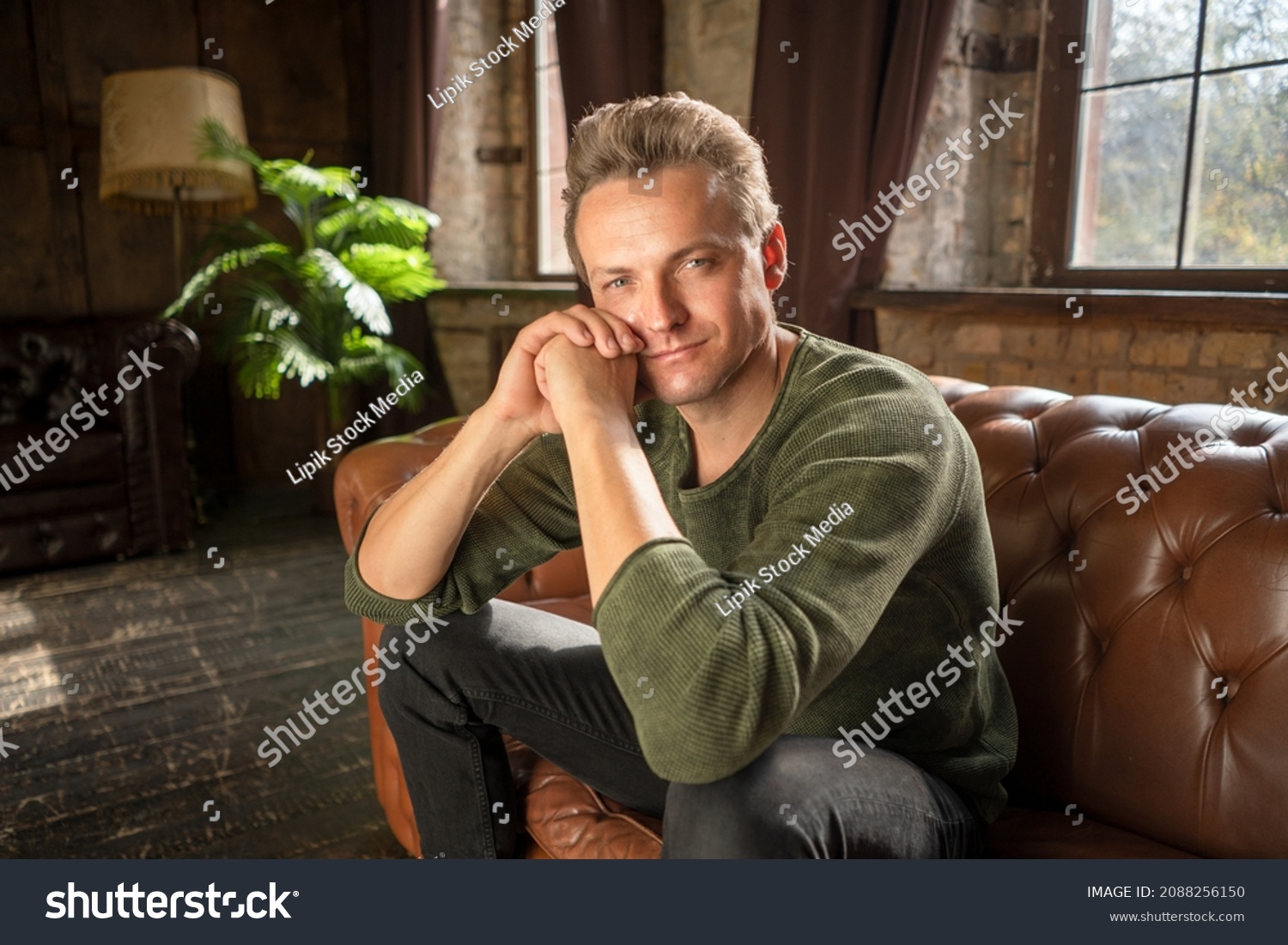 Attractive Male of 35s with Loving Eyes in Casual Clothes Sits on a Soft Leather Sofa in his Luxury Living Room of his Country House. Close-up. . High quality photo #2088256150