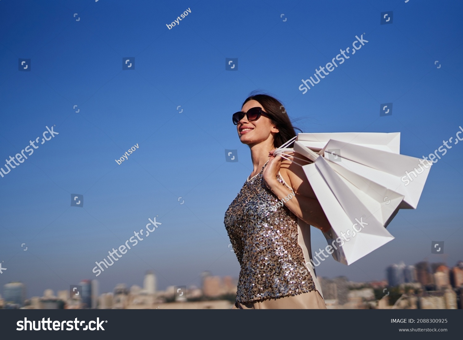 Attractive caucasian fashionable woman with white shopping bags going in front of blue sky and urban city background. Glam shopaholic brunette girl in sunglasses. Retail, consumerism or sale concept #2088300925