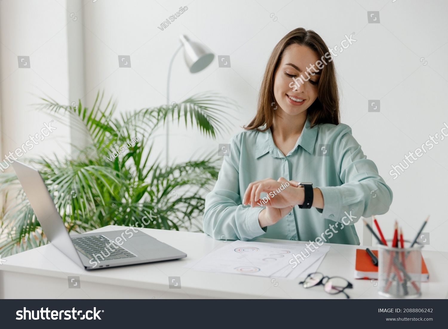 Young smiling happy successful employee business woman 20s in blue shirt look at smartwatch time sit work at workplace white desk with laptop pc computer at office indoors. Achievement career concept #2088806242