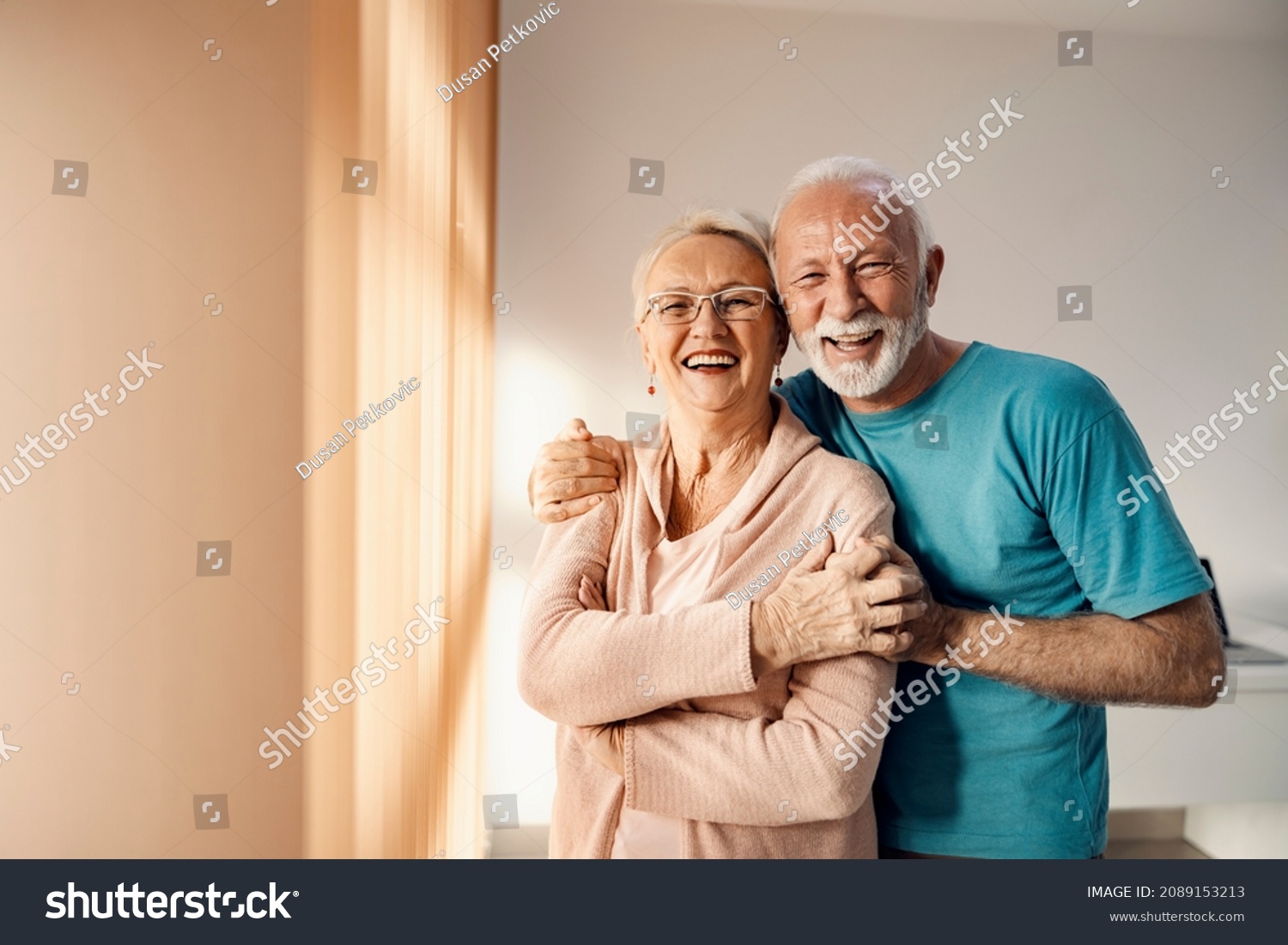 Senior couple hugging in a nursing home. A happy senior couple standing next to a window in a nursing home, hugging and smiling. They have all care they need. #2089153213