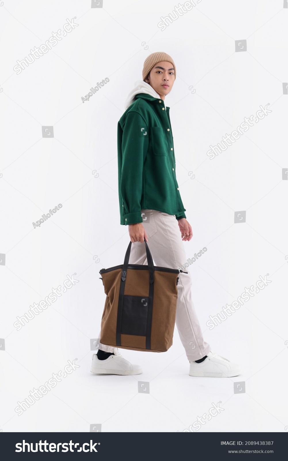 Full body young man wearing a white hoodie with green shirt with hat holding handbag walking in studio

 #2089438387