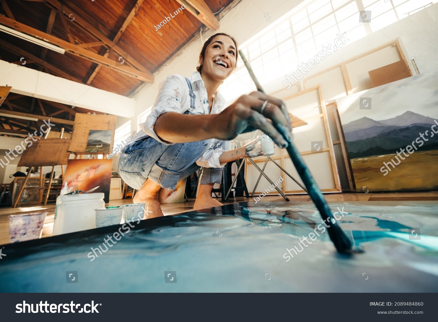 Innovative young artist painting on a large canvas in her studio. Happy female artist looking away cheerfully while working on a new artwork. Smiling woman working on the floor in her atelier. #2089484860