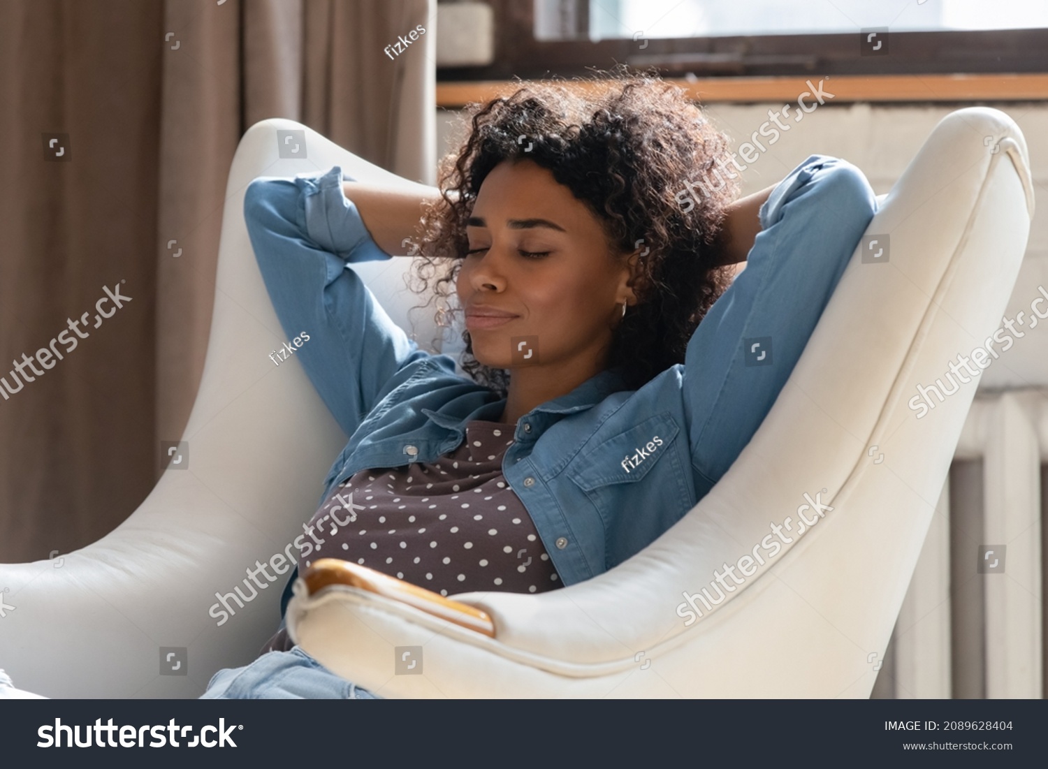 Serene African woman put hands behind head sit on leather armchair relaxing inside modern cozy air-conditioned living room. Climate control, fresh air, no stress weekend, fashionable furniture concept #2089628404