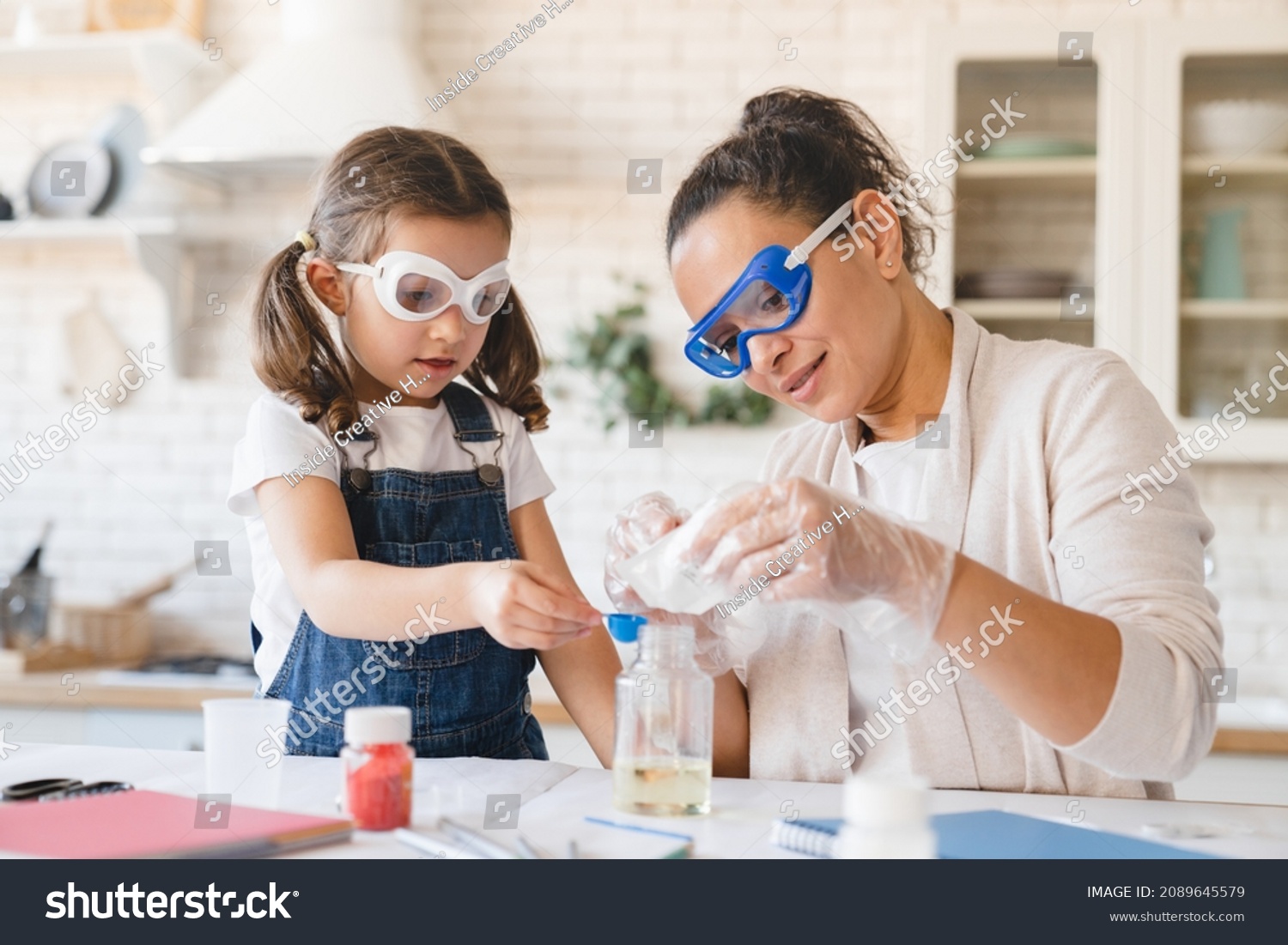 Scientific experiment at home. Laboratory tests for school homework. Parent mother with daughter kid making chemical test at home kitchen. Nanny babysitter teacher help in protective glasses #2089645579