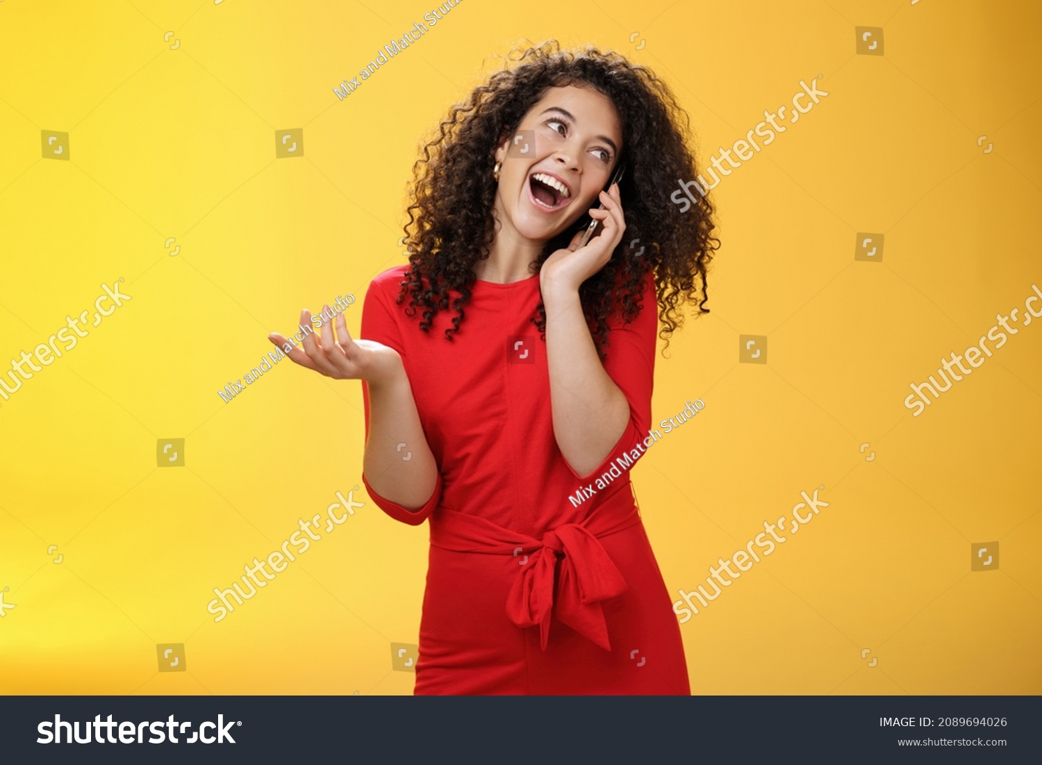 Talkative glamour silly girl with curly hair having fun feeling carefree and happy taling on mobile phone turning away as laughing joyfully gesturing with hand holding smartphone pressed to ear #2089694026