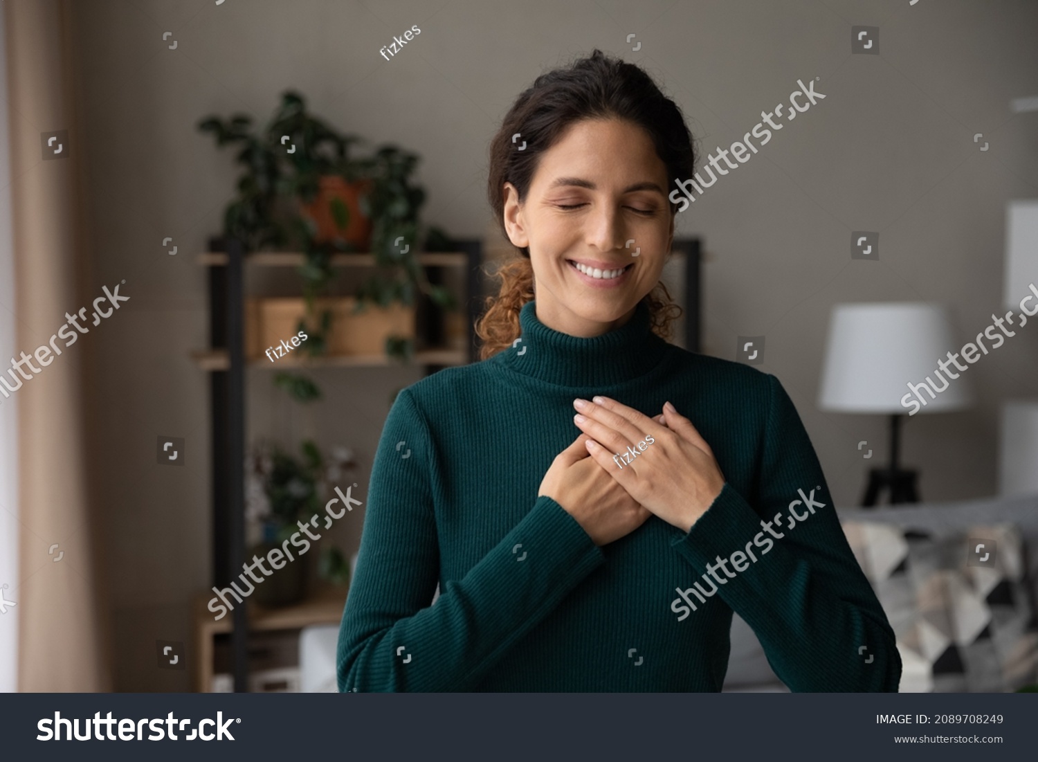 Smiling young Hispanic woman hold hands at heart chest feel grateful and thankful. Happy millennial Latino female believer show love and care support pray or mediate. Faith, superstition concept. #2089708249